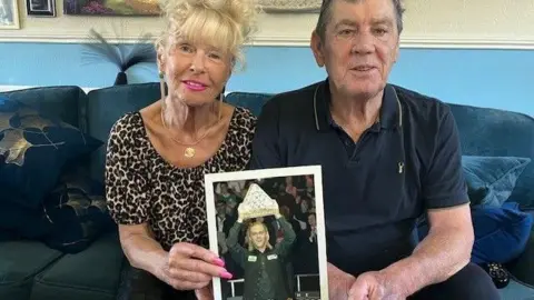 David Spereall/BBC Paul Hunter's parents hold a picture of their son lifting the Masters trophy. They are sat on a sofa and smiling into the camera. Kristina has blonde hair and is wearing a leopard-print shirt. Alan has grey hair and is wearing a dark-coloured polo shirt.