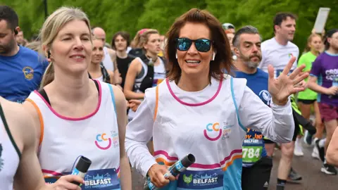 Two woman dressed in white running bibs, one of whom has brown hair, mirrored sunglasses and is waving to the camera. The other has blonde hair and has her eyes shut. 