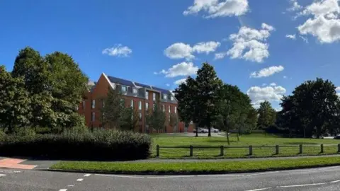 An artist's impression of the four-storey building which would house the flats. It is a red-brick building with a grey-coloured roof, positioned behind a patch of grass.