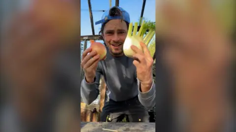 Julius Roberts holds up an onion in his right hand and a head of fennel in his left. He is wearing a grey sweatshirt and a blue baseball cap on backwards.