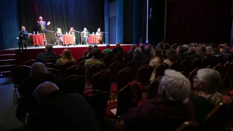 A patchy audience, with around one third of the seats empty, sits in high back chairs at players theatre Lowestoft to watch Jess Asato and other speakers on stage