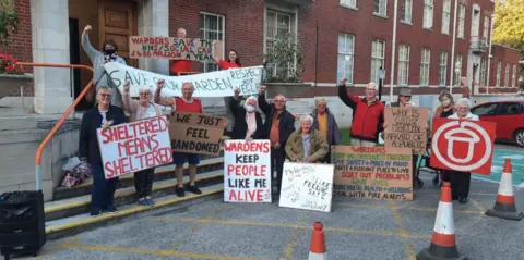 A group of people waving signs which say 'Wardens keep people like me alive' and 'sheltered means sheltered.' They are sheltered housing residents, stood in protest outside the council offices in Euclid Street in Swindon.