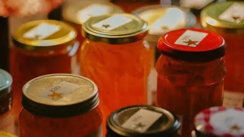 Dozens of jars of marmalade are gathered on a table. The contents are various shades of orange. All of the jar lids are labelled with a gold star.