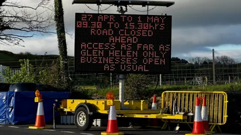 A large portable matrix sign on a yellow trailer with the details of the closure on it. There are traffic cones and crash barriers around it.