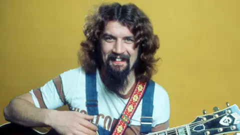 Glasgow to celebrate Sir Billy Connolly with weekend of events 1 BBC Billy Connolly in a photograph taken in the 1970s. He has long hair and a long goatee beard. He is wearing a white t-shirt and blue dungarees, and holding a guitar.