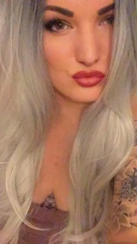 Michaela Allen A woman is facing the camera, with long, wavy, light silver‑blonde hair framing the face. The person has defined eyebrows, dark eye makeup, and is wearing dark red lipstick. 