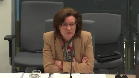 Senedd Commission Eluned Morgan being questioned in committee. She is leaning against the desk and has her arms folded.