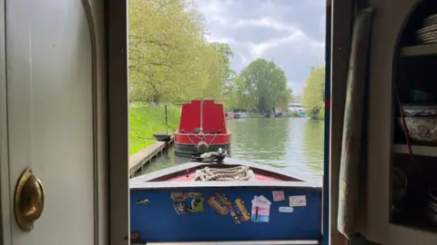 Tom Jackson/BBC The view of a river and other moored boats of different colours from inside the thin door frame of a canal boat. There are trees and grass on the edges of the canal and the river water is a murky green colour. The sun is trying to peek through a cloudy sky. The boat directly in front of where the photo is being taken is red. It has been tied to the side of the river with a rope.