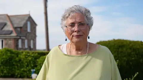 BBC/Keo Films & Button Down/Mei Williams Carole Wyatt is standing out side, looking at the camera. She has short grey hair, and is wearing glasses and a pale green t-shirt. She is standing in front of a hedge with a house in the distance.