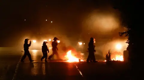 Reuters Five police officers in full protective gear, including a helmet. They are walking past two areas of fire and a police land rover can be seen in the background.
