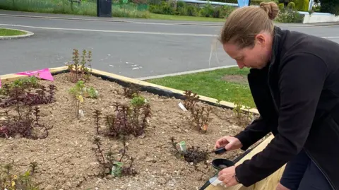 Katie Wilkinson is tending to a small herb in a large wooden planter which has others all around it. She has blonde hair tied back in a bun and she is wearing a black fleece and black shorts. There is a grassy area and a road behind her.