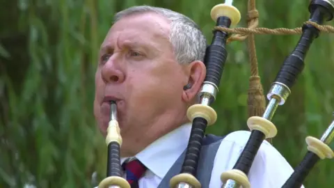 BBC A man blowing into bagpipes, he has short grey hair and is wearing a white shirt and red and blue tie. Greenery is behind him. 