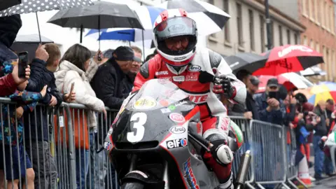 A motorbike rider, dressed in red and white protective gear, is on a racing bike with the number 3 on the front. He has a helmet on but you can tell he is smiling at the camera. There are rows of people behind him in the street. They are separated by grey metal dividers. A number of the spectators have umbrellas up.