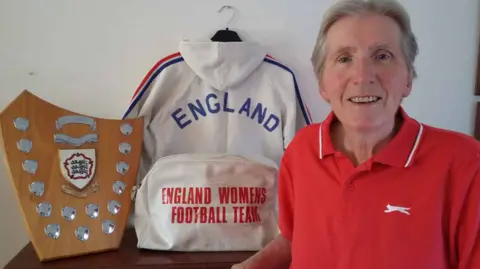 Wendy Owen smiling at the camera in a red polo shirt with her England get and trophy in the background.