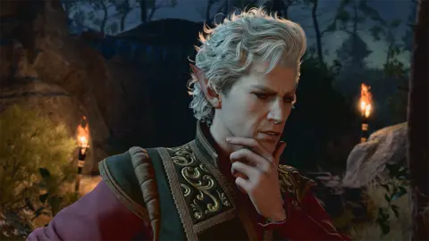 Larian Studio Screenshot shows a male character dressed in an elaborately embroidered jacket holding his chin in his hand. He looks thoughtful, but also as if he could be scheming or plotting. He appears to be in a woodland clearing lit by two flaming torches.