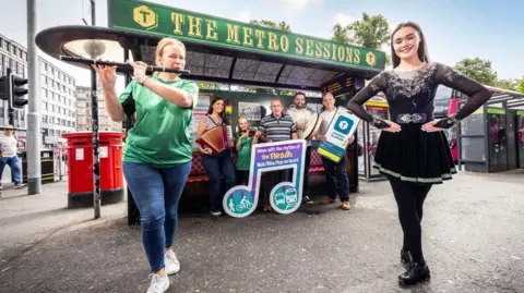 Belfast City Council Two women, one holding a flute and wearing blue jeans and a green t-shirt, the other with long brown hair and wearing a black Irish dancing dress. They are standing in front of a stall with a sign above it reading "the metro sessions". Three men, a woman and a child and standing under the stall. 