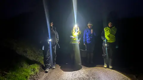 Four people, two in reflective jackets, all carrying torches on a narrow lane at night