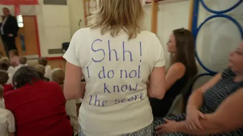 A teacher wearing a white tee-shirt. On the back is the message: Shh, I do not know the score.