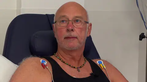 Patient Andy Mouncey sat on a chair looking away from the camera. He has some medical equipment attached to each of his shoulders. 