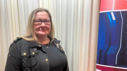 Amy Holmes/BBC A woman wearing a black jacket and dark top stands in front of light-coloured curtains, next to a display panel featuring bold red and blue graphics and images of screens.