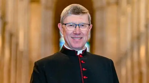 Keith Heppel The Very Reverend Mark Bonney has short brown hair. He is wearing black framed glasses and a black and red cassock.