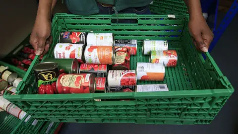 PA Media Close up of a green plastic basket with tins of tomatoes in it being held by a foodbank volunteer