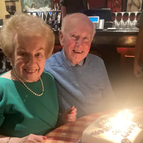 Joe Connolly with his wife. They are sitting in a restaurant with a cake in front of them - there's several candles on it. Joe is wearing a blue jumper and his wife is wearing a green top with a string of pearls. Both are smiling at the camera.