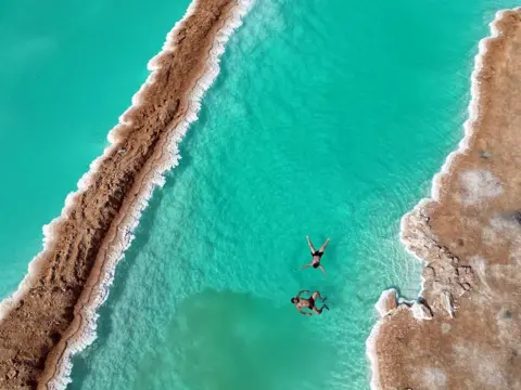 FAREED KOTB / ANADOLU / GETTY IMAGES Two men in swimming shorts float on turquoise-colour waters. The banks have salt-encrusted edges. 