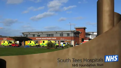 LDRS The outside of the 'Regional Major Trauma Centre'. In the foreground is a curved brown wall with South Tees Hospitals NHS Foundation Trust written on it. Multiple ambulances can be seen outside the building.