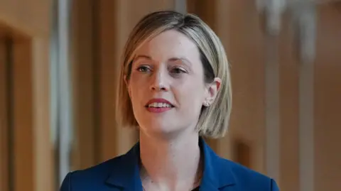 Jenny Gilruth has a blonde bob and looks off to one side. She wears a black top and blue blazer