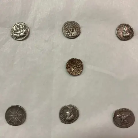 Seven old silver coins lie on a white background. The name "Caesar" can be read on one of them.