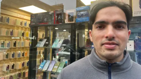 Moby is looking at the camera and is standing outside his shop. He has black hair and a grey fleece on. Through the window behind him are mobile phones and mobile phone covers on shelves.