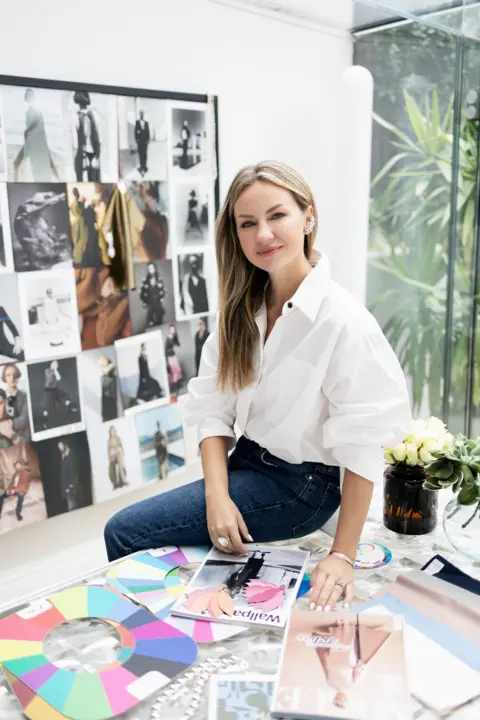 Oriona Robb Image of a blond woman sitting at a table with magazine and colour wheels on the table and a mood board behind her of models wearing different outfits