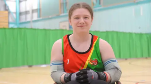 Sarah Burrows, captain of Penrith Pumas wheelchair rugby team. She has light brown hair tied back and blue eyes. She is inside a sports hall and is wearing a red, yellow and green vest and protective sport equipment including gloves and elbow pads.
