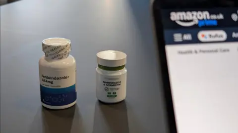 Two white pill bottles on a table, one labelled Fenbendazole and the other Ivermectin. A laptop stands beside them, with the Amazon logo visible on the screen. 