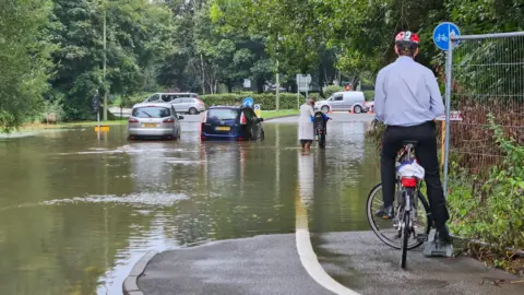 Anna J A flooded road with two cars stuck in the middle of it. A man on a bike is surveying the scene on a pavement above the water. Another woman is trying to push her bike through the flooded road