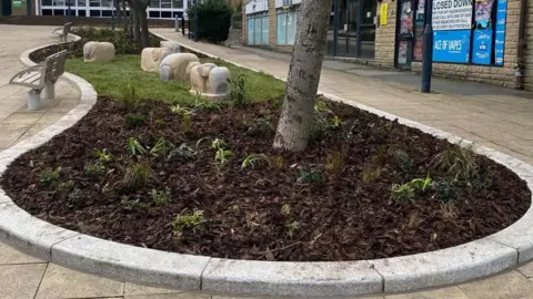 Dan Jones A small paved square contains a circular planted bed filled with mulch, young plants and a tree, surrounded by curved stone benches and sculptural stone shapes. Shops and closed storefronts line the background, giving the space an urban, slightly quiet feel.