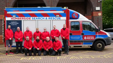 A sideways view of a van with sliding doors on the side. Along the top are the words Humberside Search and Rescue. In front of the van are 10 men and one woman all wearing bright red, windproof jackets. 
