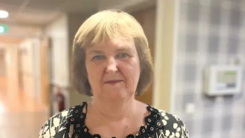 Linda Graham is looking directly at the camera. She has a brown/blonde bob and blue eyes. She is wearing a black and cream patterned top which has a scalloped neck line.