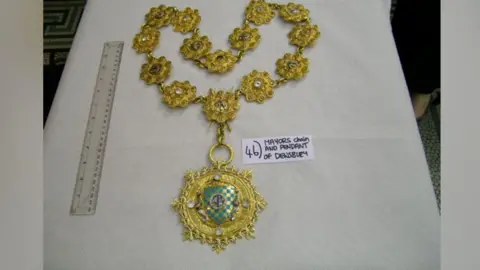 A gold-coloured mayoral chain designed to fit around someone's neck and torso. A green broach is in the middle of the circular pendant. A 30cm ruler sits to one side of the chain. A small piece of paper sits on the other side with the words 'Mayors (sic) chain and pendant of Dewsbury'. 