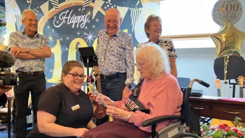 A woman in a black top smiles at a woman in a wheelchair who is looking at a card. People holding musical instruments are standing behind her and smiling. There is a banner behind her, as well as balloons.