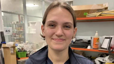 Who are the workers who will fire Scotland's space industry? 1 BBC Millie Brown, a young lady with brown hair in a ponytail, smiles from inside an engineering lab at her university. There are busy-looking shelves and piles of equipment, boxes and other items behind her.
