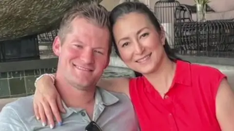Close up of Blair and Tina Campbell smiling at the camera. She has her arm round his shoulders and is wearing a red top with her hair tied back. He is wearing a pale grey shirt and has short hair spiked up on top