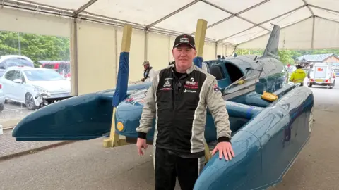 Dave Warby standing in front of Bluebird in the workshop tent. He is wearing a balck cap and black-and-grey overalls.