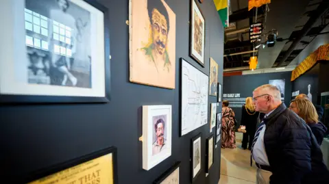 Hopetown Darlington Two people can be seen looking at an exhibition wall lined with pictures and newspaper clippings, celebrathing Arthur Wharton's achievements. The rest of the exhibition can be seen behind them, and other visitors.