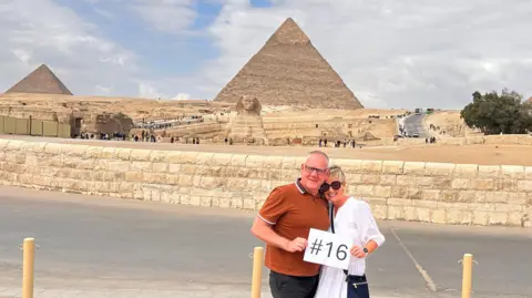 Tony and Tracy Raine The couple in front of the Great Pyramids in Egypt. Tony has a brown t-shirt on and Tracy has a white blouse with sunglasses. They are holding an A4 sheet of paper with the number 16 printed.