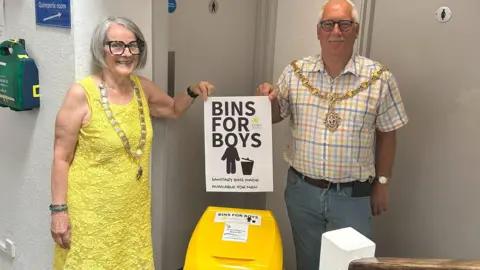 Liskeard Mayor Cllr David Braithwaite and Deputy Mayor Cllr Sylvia Berry are stood outside a toilet facility holding a Bins for Boys poster above a bright yellow bin. They are both looking straight at the camera and are smiling. 