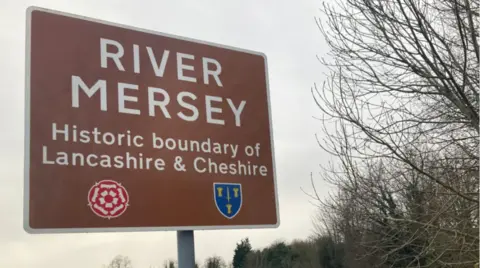 A sign next to the River Mersey in Greater Manchester which reads "River Mersey, historic boundary of Lancashire and Cheshire"