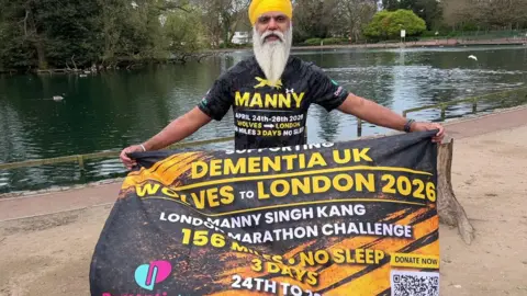 Manny Singh Kang - a man with a long grey beard and yellow turban stands next to a lake in West Park, Wolverhampton holding a large banner with writing on it