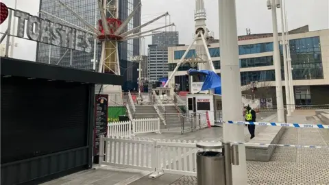 A view of the fairground ride with a police officer standing beside some police tape - a tall central metal structure with swings flying around in a circle.